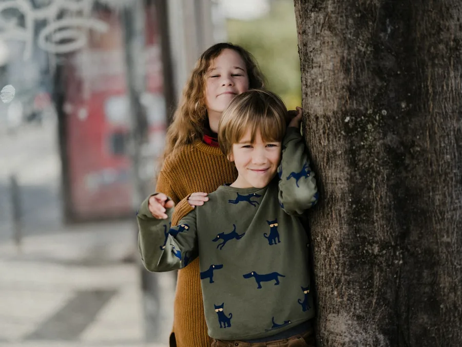 Two kids leaning against a tree trunk on a city sidewalk, smiling